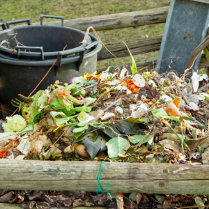 A vibrant compost heap overflows with a variety of organic waste against an outdoor backdrop. Leaves, vegetable peels, and other green waste create a colorful mosaic atop the wooden edges of a compost bin. In the background, a black bucket and a metal shovel await use, signaling ongoing gardening activities