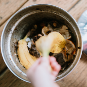 "Overhead view of a metal bowl filled with kitchen compost, including banana peels, coffee grounds, and vegetable scraps. A hand holds a fresh piece of vegetable waste above the bowl, ready to drop it into the mix. The bowl sits on a wooden surface, illustrating a step in the eco-friendly practice of composting food waste.