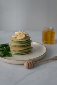 spinach pancakes in a stack with sliced bananas on top and a few few fresh leaves for decoraion with a jar of honey and a drizzle stick 