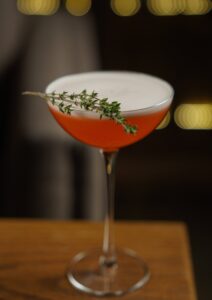  "An elegant cocktail with a rich amber hue, garnished with a sprig of thyme, set against a backdrop of warm bokeh lights."