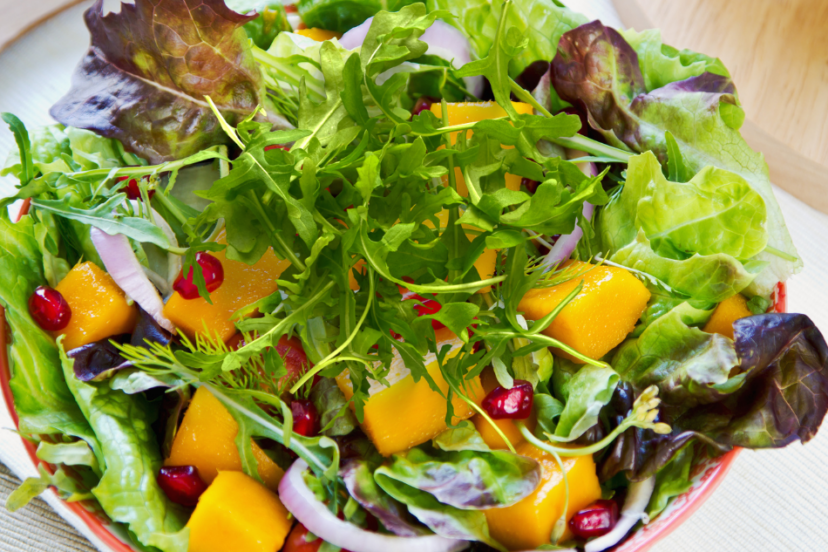A vibrant bowl of mixed leaf salad featuring fresh arugula, colorful lettuce, succulent mango chunks, and juicy pomegranate seeds.