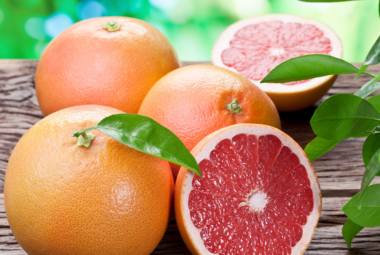 Whole and halved grapefruits with rich, ruby-red flesh and green leaves, resting on a wooden surface.