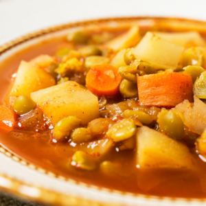 A close-up of a bowl of mixed vegetable soup, rich in color with potatoes, carrots, and peas in a tomato-based broth.