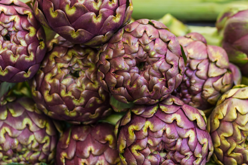 "Close-up of a pile of vibrant purple artichokes with green highlights, freshly harvested and displayed at a market."