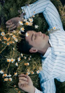 Article: Chamomile Companion Plant. Pic - A serene scene of a young man lying in a meadow, eyes closed, surrounded by white camomile flowers with a few yellow centers, and green foliage.