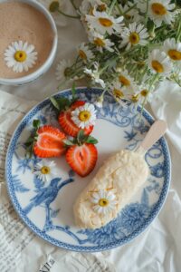 Article: Chamomile Companion Plant. Pic - A beautifully arranged breakfast setting featuring a blue and white porcelain plate with a vanilla ice cream bar, fresh strawberry halves, and camomile flowers, next to a cup of frothy coffee and a bouquet of camomiles on a text-covered linen.
