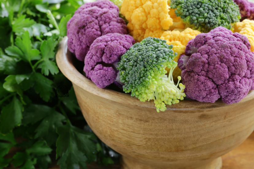 A wooden bowl filled with vibrant, colorful cauliflower florets in purple, yellow, and green, placed on a wooden table with fresh parsley in the background.