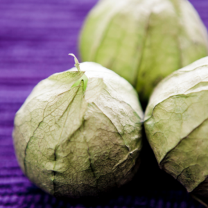 Article: Tomatillo Companion Plants . Pic - "A trio of green tomatillos wrapped in their natural, dry husks, set against a vibrant purple background."