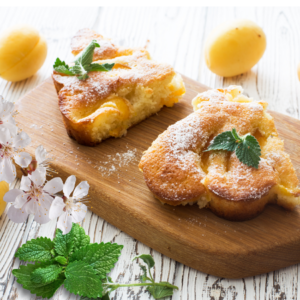 Slices of apricot cake on a wooden board, garnished with mint leaves and apricot blossoms, with whole apricots in the background.