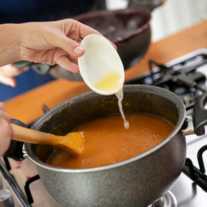  A person adding oil to a simmering pot of apricot jam on a stove, stirring with a wooden spoon.