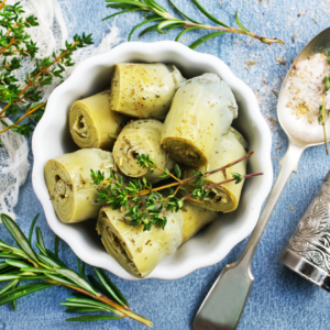 Article: Artichoke Companion Plants. Pic - Marinated artichoke hearts in a white ceramic bowl garnished with fresh thyme, surrounded by herbs on a blue textured background.