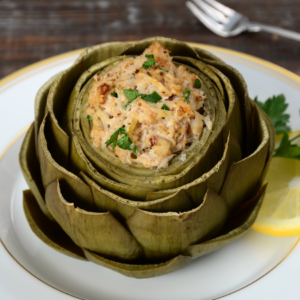 Article: Artichoke Companion Plants. Pic - A cooked artichoke on a plate, its center filled with a creamy crab stuffing, garnished with parsley and lemon on the side.