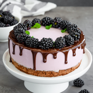  A blackberry cheesecake topped with fresh blackberries, drizzled chocolate, and mint leaves on a white cake stand.
