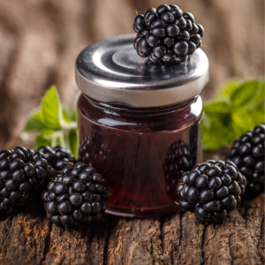 A small jar of homemade blackberry jam topped with a blackberry, surrounded by more blackberries and mint leaves on a rustic wooden surface.