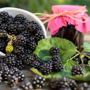 A bowl overflowing with ripe blackberries, next to a jam jar and under a pink flower, on a wooden surface.