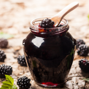 Article:Blackberry Companion Plants. Pic - A jar of homemade blackberry jam on a rustic wooden surface, with a spoonful of jam and scattered blackberries around.