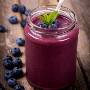 A glass jar of smooth blueberry smoothie topped with fresh blueberries and a mint leaf, on a rustic wooden table.