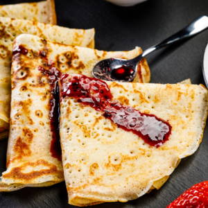 Golden crepes on a plate, drizzled with blueberry jam, served with a spoonful of jam on the side on a black surface.