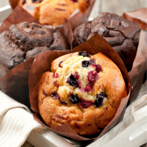 Article: Companion Planting Blueberries. Pic -Close-up of a freshly baked blueberry muffin in a brown paper cup, surrounded by other muffins in a wooden tray.
