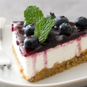 Article: Companion Planting Blueberries. Pic -A slice of blueberry cheesecake with a glistening top layer of blueberry glaze, fresh blueberries, and a mint leaf on a white plate.