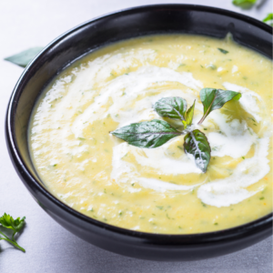 Creamy broccoli soup in a black bowl garnished with fresh basil leaves, served on a light-colored table.