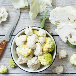 A bowl of cauliflower florets on a wooden table, surrounded by a knife and cauliflower pieces.