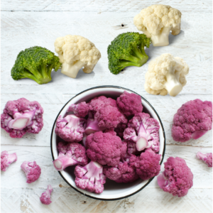 Article: Cauliflower companion planting. Pic - A bowl of purple cauliflower florets on a white wooden table, surrounded by pieces of white cauliflower and broccoli.
