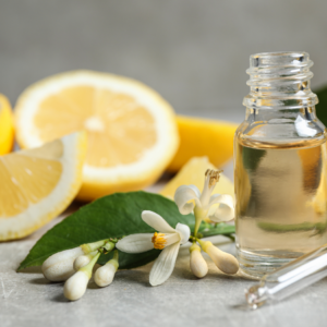 A small bottle of essential oil next to lemon slices and citrus blossoms on a gray surface.