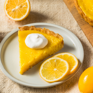  A slice of lemon tart topped with a dollop of whipped cream, accompanied by fresh lemon slices on a light ceramic plate.