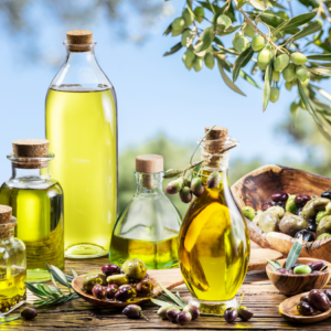 Various bottles of olive oil with cork stoppers, surrounded by fresh and marinated olives, set outdoors with olive trees in the background.