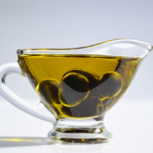 A glass jug filled with golden olive oil, with whole olives submerged inside, set against a white background.