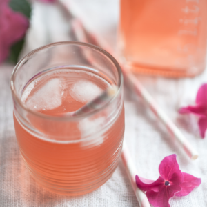 Article: Rhubarb Companion Plants. Pic - Refreshing rhubarb cocktail served in a glass with ice cubes, accompanied by a clear glass bottle and pink flowers on a linen tablecloth.