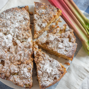 Article: Rhubarb Companion Plants. Pic - Freshly baked rhubarb pie dusted with powdered sugar, served on a white plate with fresh rhubarb stalks beside it on a blue napkin.