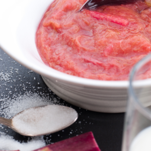 : A bowl of stewed rhubarb next to a spoonful of sugar and a glass of water.