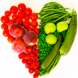 An assortment of fruits and vegetables arranged in a heart shape, including tomatoes, peaches, strawberries, green beans, peas, apples, and zucchinis, on a white background.