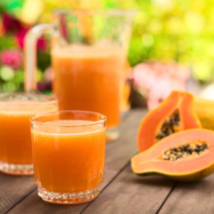 Article: Papaya Enzymes for digestion. Pic - A pitcher and two glasses of fresh papaya juice with halved papayas on a wooden table in a garden setting.