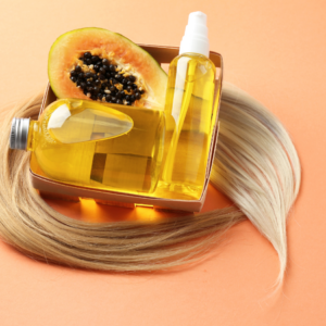 Article: Papaya Enzymes for digestion. Pic - A set of bottles containing papaya oil is displayed with a halved papaya showing seeds in the background and a strand of blonde hair. The bottles, filled with golden-yellow oil, are placed on an orange surface.