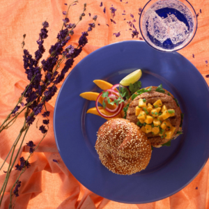 Article: Papaya Enzymes for digestion. Pic  - A sesame seed burger bun with a patty topped with diced papaya salsa, accompanied by mango slices, red onion rings, and a lime wedge on a blue plate, with a glass of sparkling water and purple flowers on an orange tablecloth.