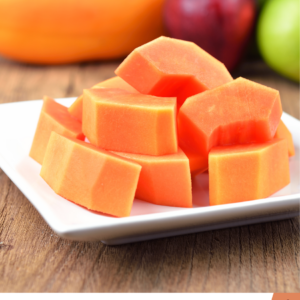 A white plate with neatly arranged cubes of fresh papaya, set on a wooden table with various fruits blurred in the background.