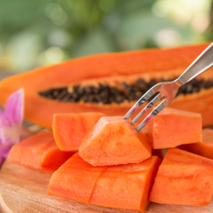 A close-up of a fork picking up a cube of fresh papaya from a wooden board, with a halved papaya showing seeds in the background.