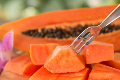 A close-up of a fork picking up a cube of fresh papaya from a wooden board, with a halved papaya showing seeds in the background.
