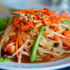 A close-up of a vibrant papaya salad with shredded green papaya, carrots, green beans, tomatoes, and peanuts, served on a white plate.