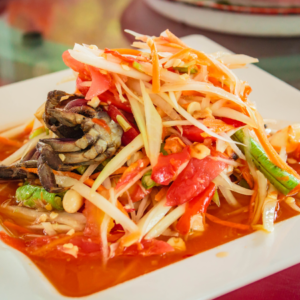 A close-up of a vibrant papaya salad with shredded green papaya, tomatoes, green beans, peanuts, and crab, served on a white plate.