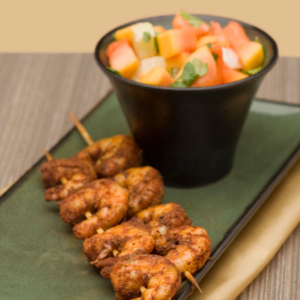 A dish featuring grilled shrimp skewers alongside a black bowl of colorful papaya salsa on a green plate.