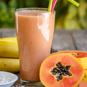 A tall glass of papaya smoothie with a couple of straws, surrounded by halved papayas, bananas, and a bowl of oats, set on a wooden surface.